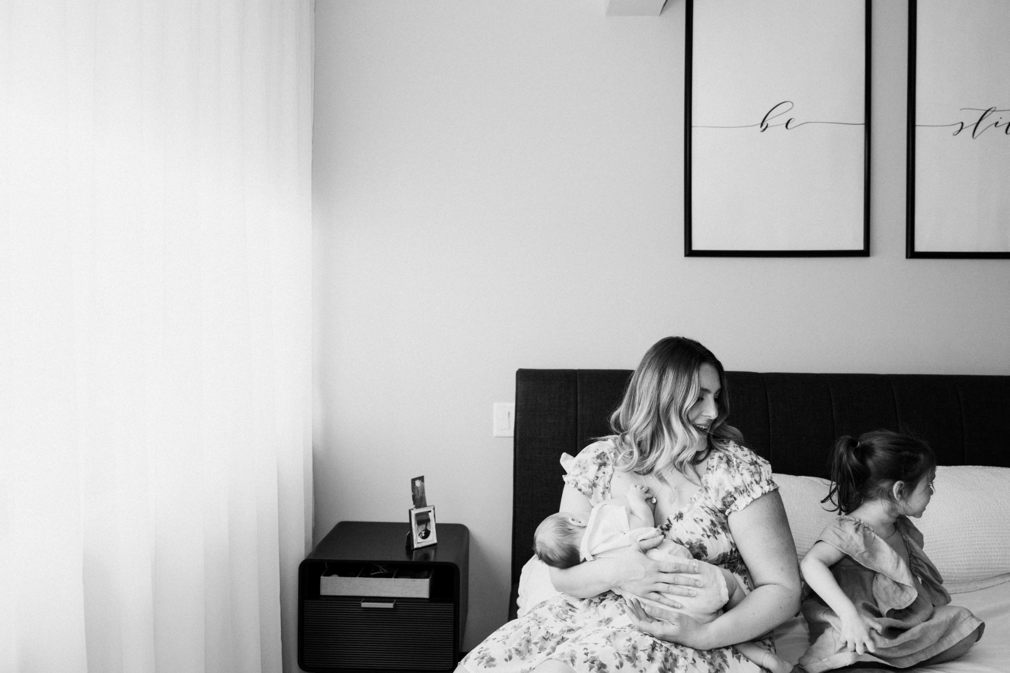 A mother nursing her newborn on the bed while her toddler sits beside her